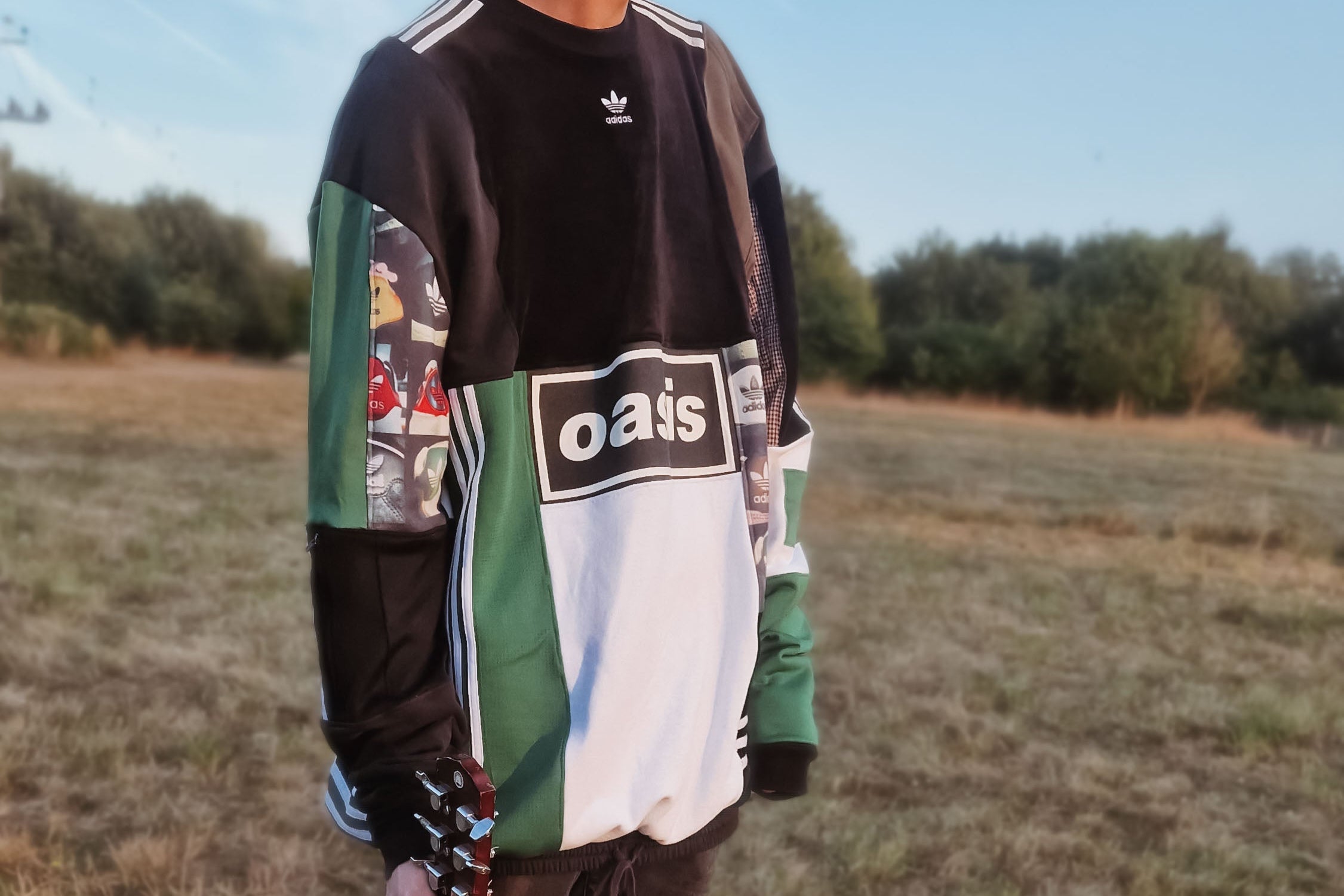 Person holding a guitar in a field wearing an upcycled jumper with 'oasis' adidas branding.